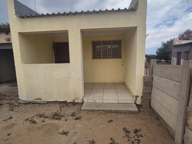Front view of a small residential structure featuring a tiled patio, security gate, and precast boundary walling in need of maintenance.