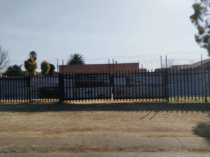 A vacant residential plot secured by metal fencing, razor wire, and a double gate, viewed from the street.