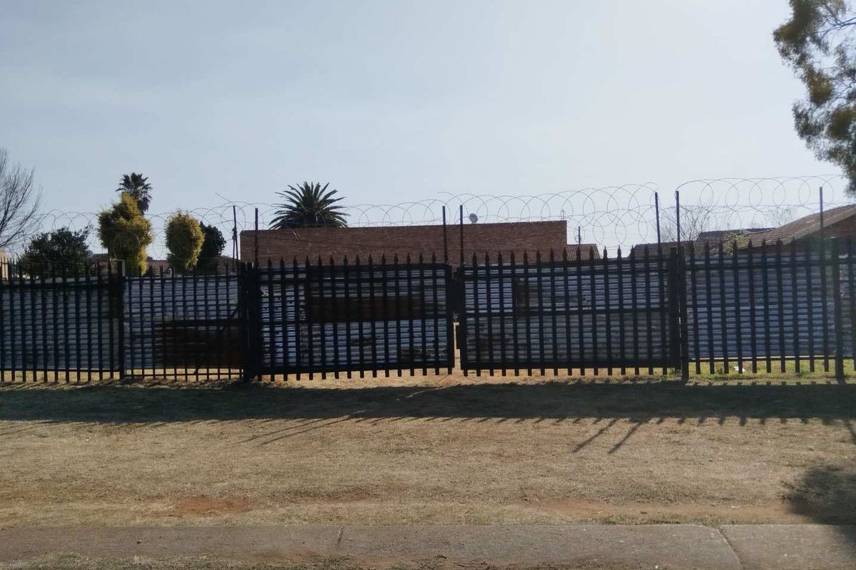 A vacant residential plot secured by metal fencing, razor wire, and a double gate, viewed from the street.