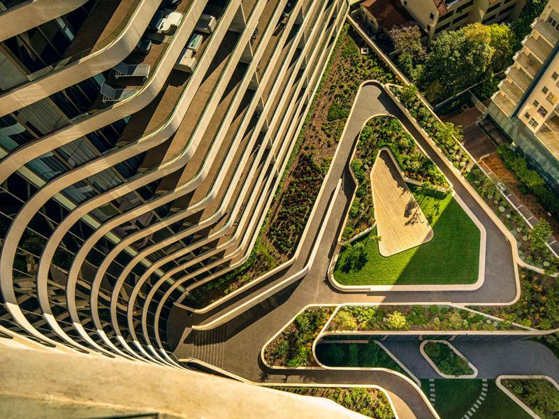 High-angle aerial view of the modern penthouse building exterior, showcasing the curved architectural facade and the landscaped ground-level garden and pathways.