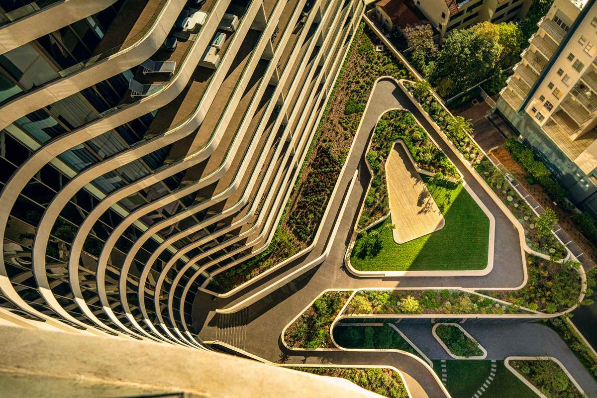 High-angle aerial view of the modern penthouse building exterior, showcasing the curved architectural facade and the landscaped ground-level garden and pathways.