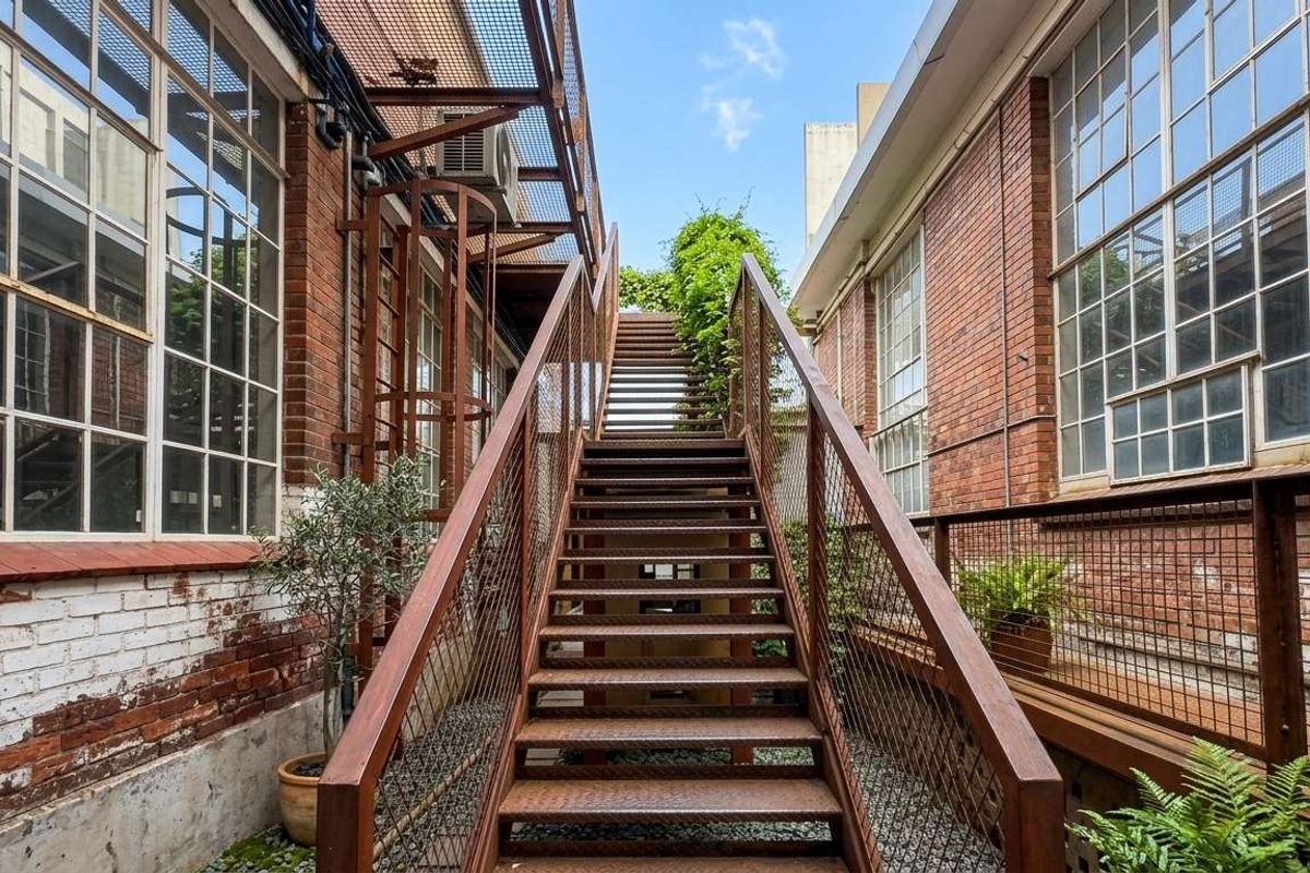 An industrial-style exterior rear courtyard featuring a prominent metal staircase, exposed brick walls, and large multi-pane windows.