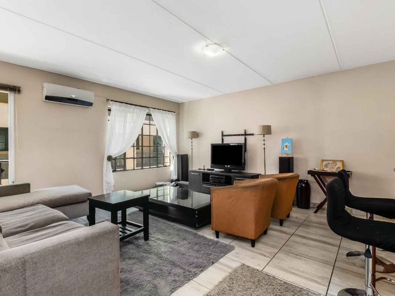 A comfortable living room featuring tiled flooring, a wall-mounted air conditioning unit, and large windows with security bars, offering a functional and well-lit space.