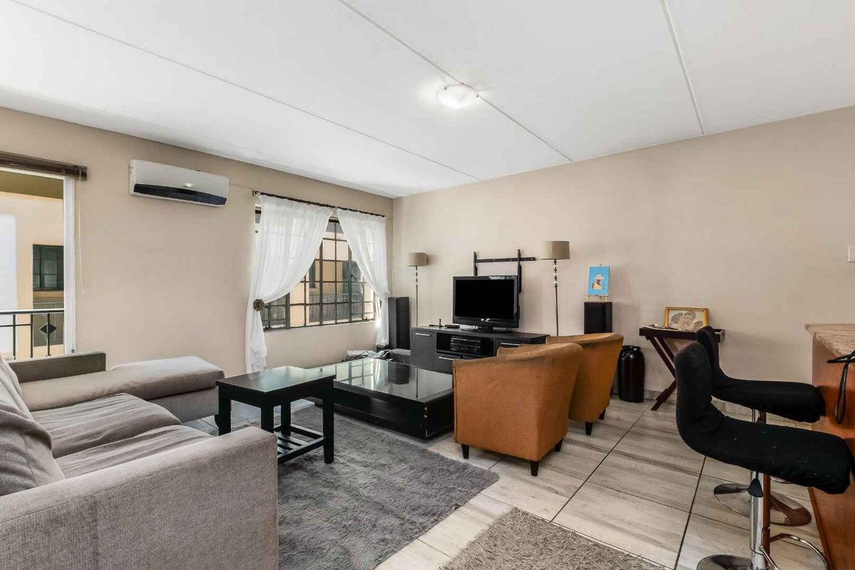 A comfortable living room featuring tiled flooring, a wall-mounted air conditioning unit, and large windows with security bars, offering a functional and well-lit space.