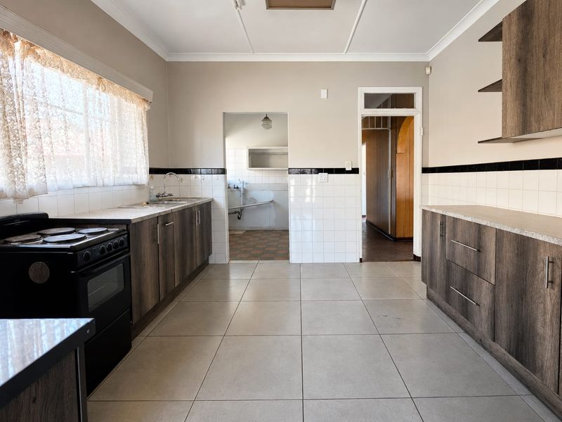 A functional kitchen featuring wood-look cabinetry, tiled backsplash, and ceramic flooring, with an adjacent scullery area visible through the doorway.