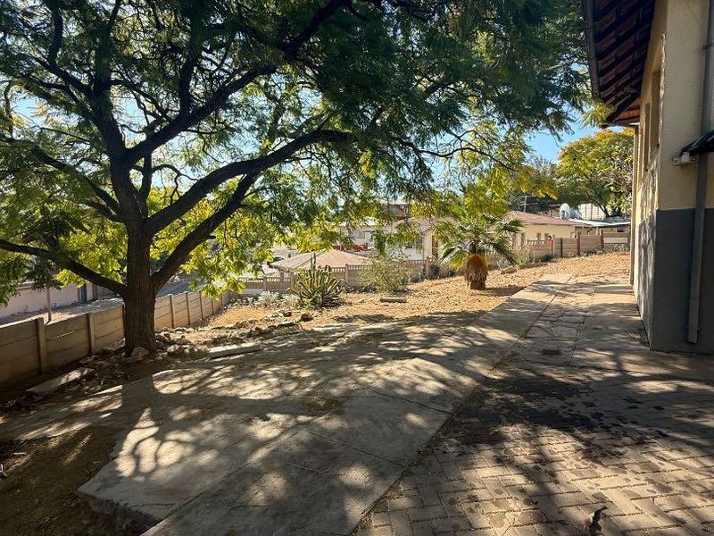 Rear exterior view featuring a paved walkway, concrete patio area, and established trees along the property boundary wall.
