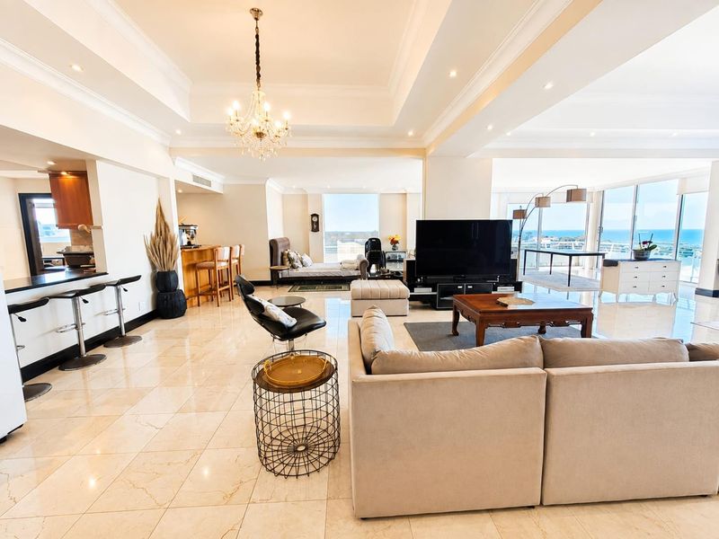 Spacious open-plan living area featuring polished marble flooring, a tray ceiling with recessed lighting, and expansive glass doors offering panoramic ocean views.