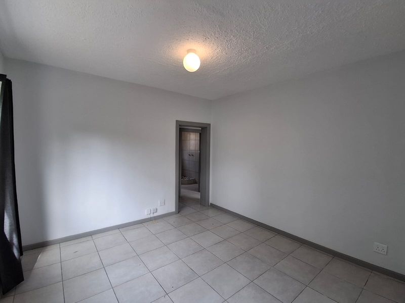 A clean, empty bedroom featuring neutral tiled flooring, grey skirting boards, and a simple ceiling light fixture, with an adjacent view into a bathroom.