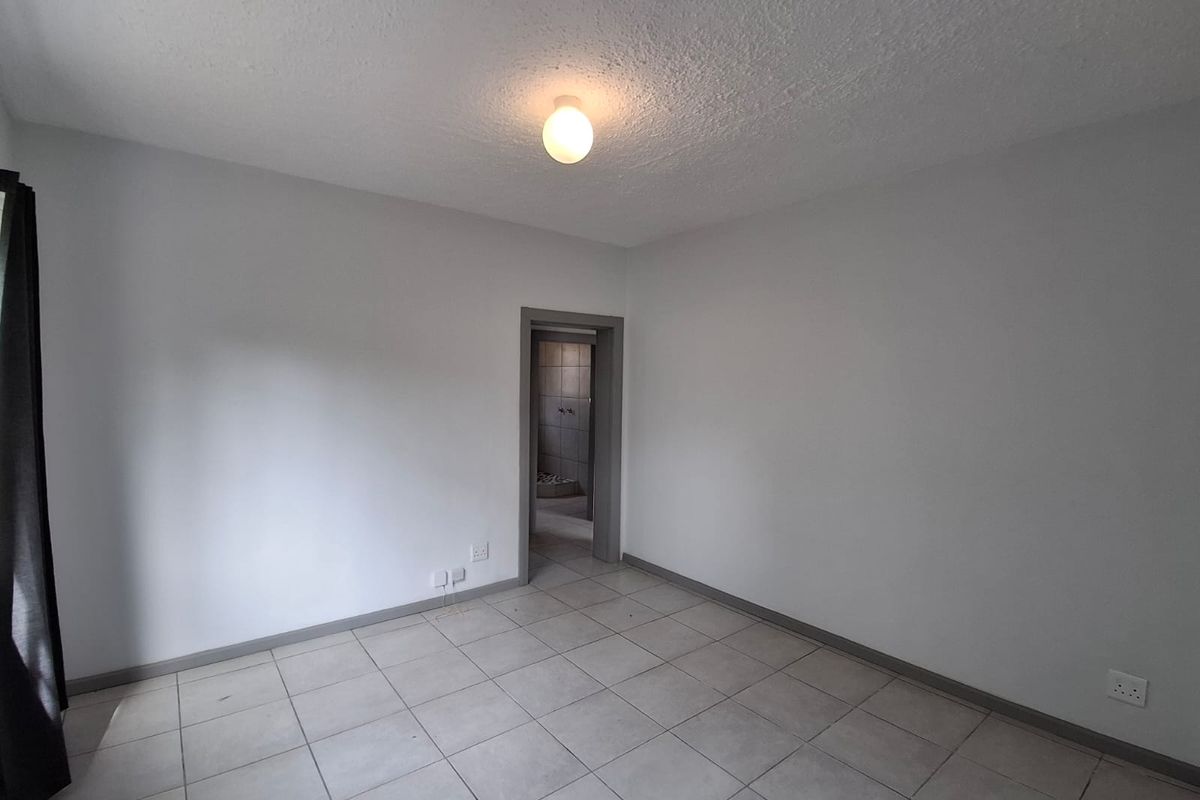A clean, empty bedroom featuring neutral tiled flooring, grey skirting boards, and a simple ceiling light fixture, with an adjacent view into a bathroom.