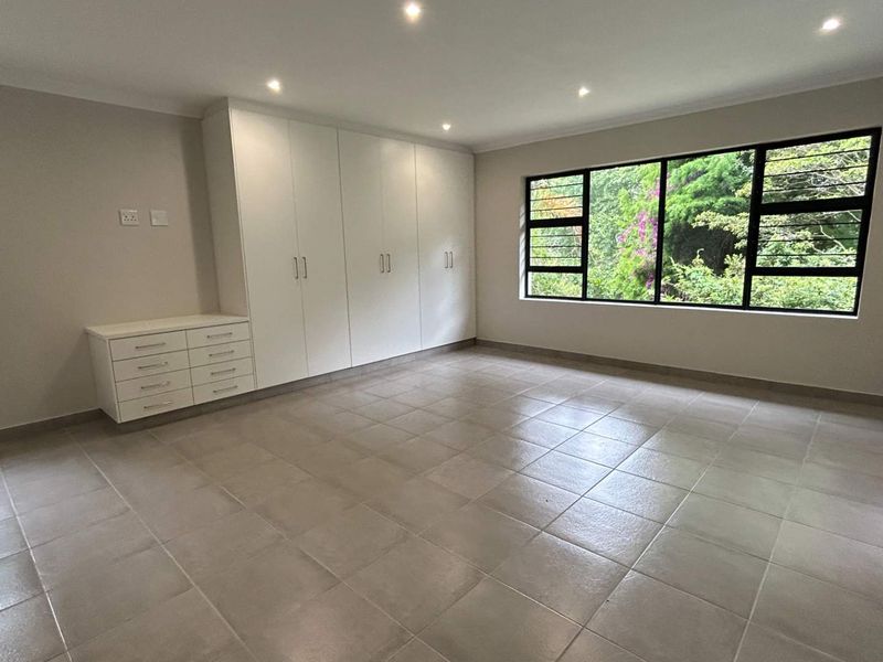 A spacious, empty bedroom featuring neutral tiled flooring, built-in cupboards, and large windows with views of the surrounding greenery.