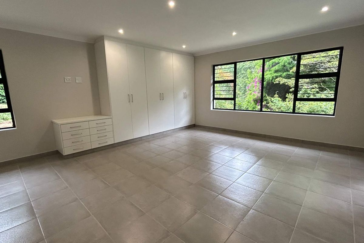 A spacious, empty bedroom featuring neutral tiled flooring, built-in cupboards, and large windows with views of the surrounding greenery.