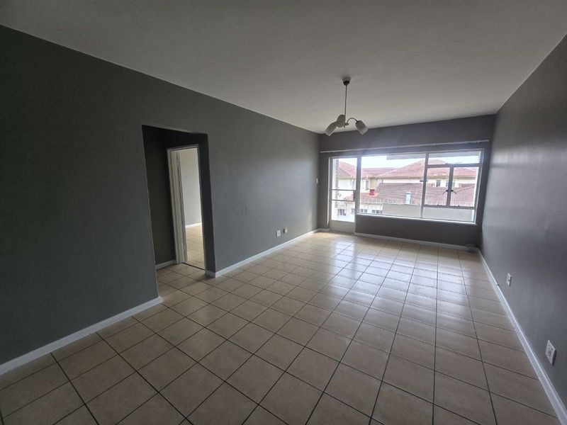 A spacious, empty living room featuring neutral grey walls, tiled flooring, and a large window providing natural light.