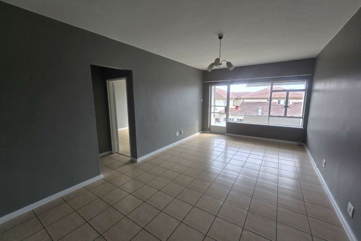 A spacious, empty living room featuring neutral grey walls, tiled flooring, and a large window providing natural light.