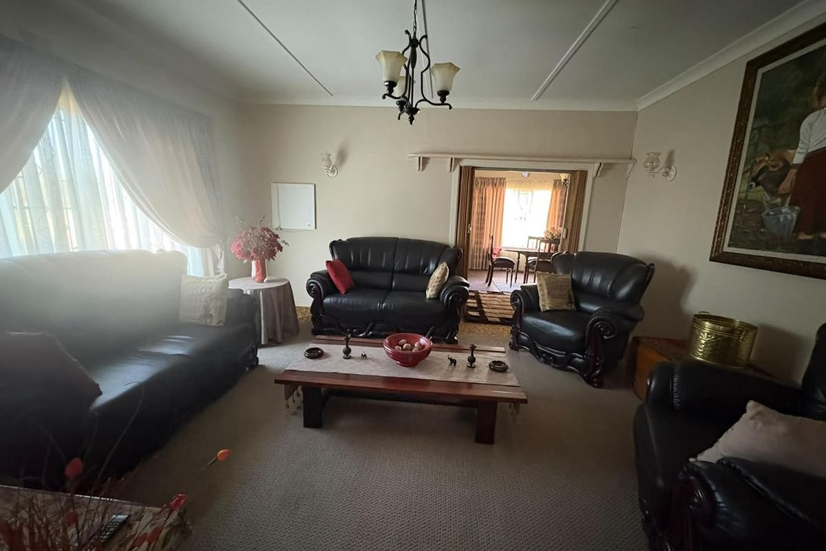 A traditional living room featuring carpeted flooring, a central chandelier, and wall sconces, with an adjacent dining area visible through a sliding door.