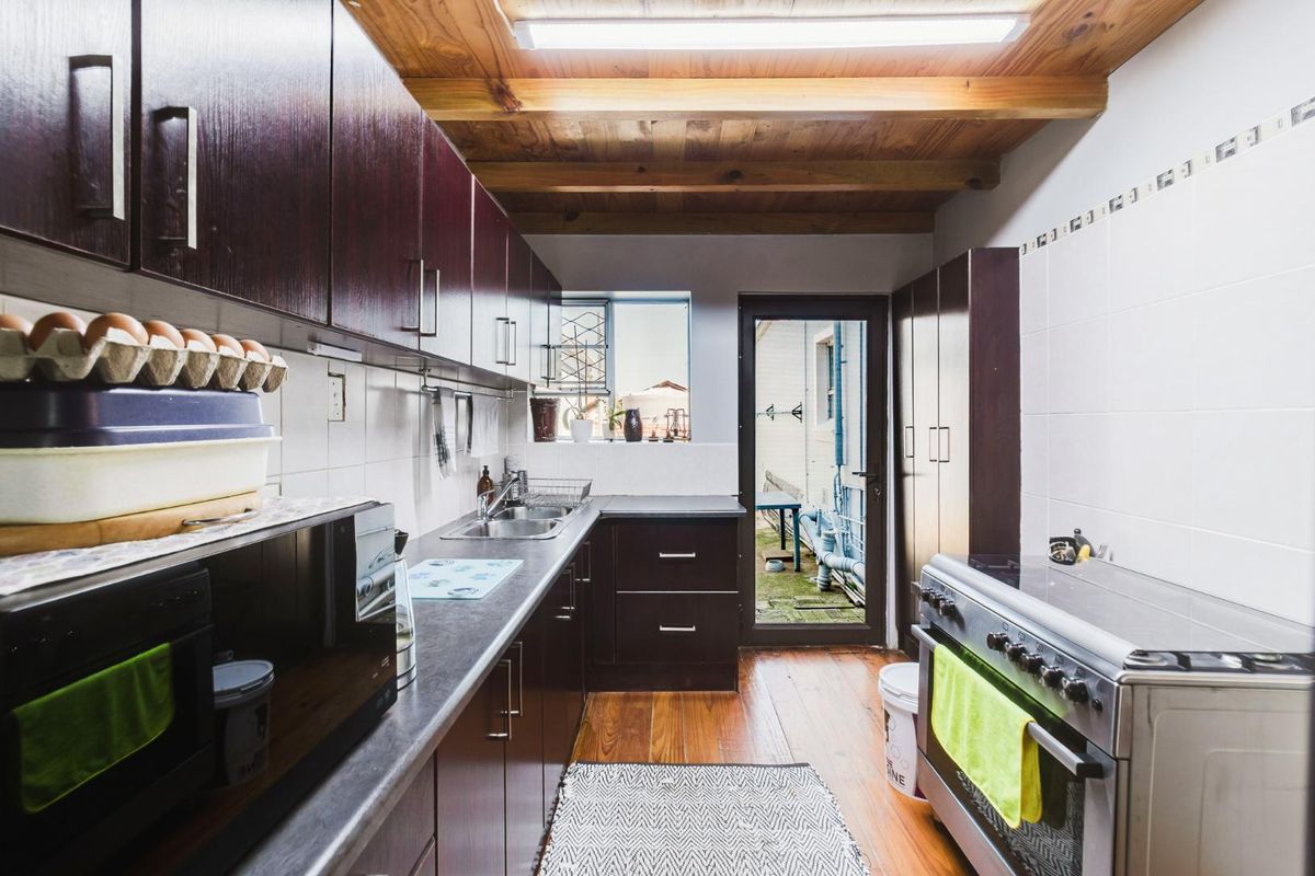 Functional kitchen featuring dark wood cabinetry, laminate countertops, stainless steel sink, and exposed timber ceiling beams with direct access to the rear exterior.