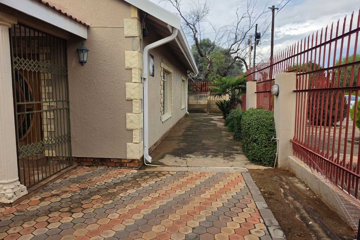 Side exterior view of the property featuring paved walkways, security fencing, and established garden borders along the building facade.