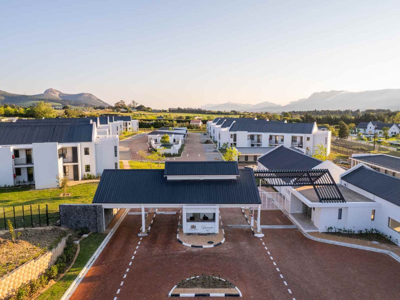 Aerial view of a modern residential apartment complex featuring a secure gated entrance, paved driveway, and well-maintained landscaped grounds with mountain views.