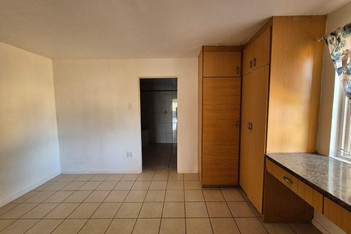 A vacant bedroom featuring built-in wooden cupboards, a matching built-in desk, and neutral tiled flooring, with direct access to an adjoining bathroom.
