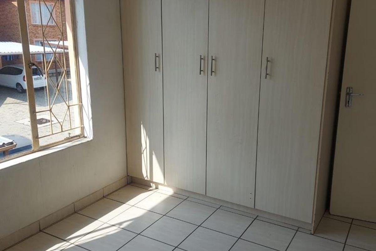 A bright, empty bedroom featuring built-in cupboards, tiled flooring, and a window with security bars.
