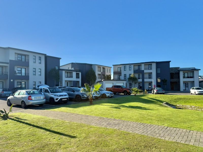 A view of the townhouse complex exterior featuring multi-story residential buildings, paved parking areas, and well-maintained communal lawn spaces.