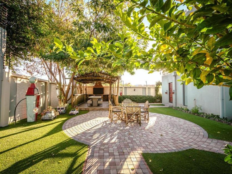 A well-maintained garden featuring a circular brick-paved patio, artificial lawn, and a covered thatched lapa with a built-in braai area.