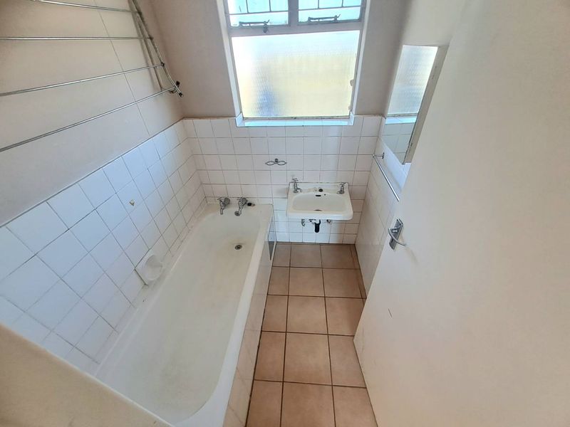 A compact bathroom featuring a standard bathtub, wall-mounted basin, and tiled finishes, requiring cosmetic updates to improve its overall condition.
