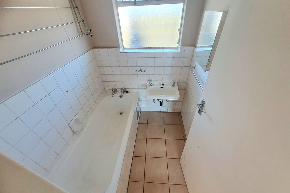 A compact bathroom featuring a standard bathtub, wall-mounted basin, and tiled finishes, requiring cosmetic updates to improve its overall condition.