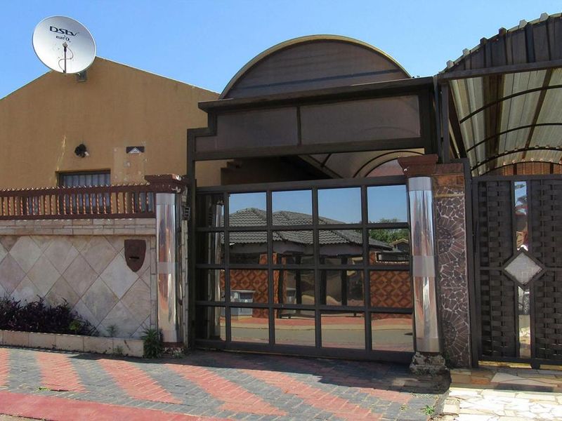 Front exterior view of the property featuring a secure sliding gate, paved driveway, and boundary wall with integrated carport structure.