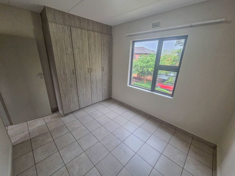 A clean, empty bedroom featuring neutral tiled flooring, built-in wooden cupboards, and a large window providing natural light.