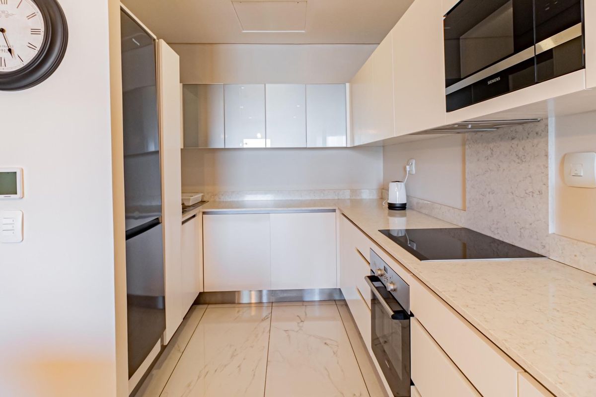 Modern galley-style kitchen featuring sleek white cabinetry, stone-look countertops, integrated oven, induction hob, and built-in microwave with polished marble-effect floor tiles.
