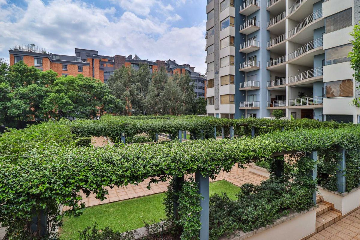 A well-maintained communal garden area featuring a large pergola, paved pathways, and manicured lawns situated between apartment buildings.