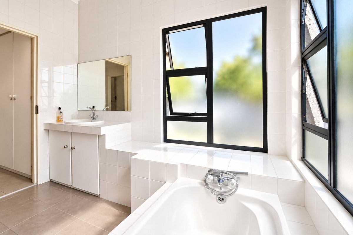 A bright and clean bathroom featuring a built-in vanity, a drop-in bathtub, and large windows with black frames, finished with neutral wall and floor tiles.