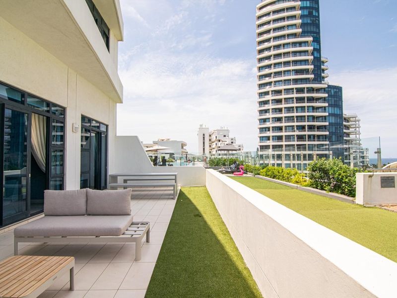 Spacious outdoor balcony featuring tiled flooring, artificial turf, and a modern glass balustrade with views of the surrounding high-rise buildings.