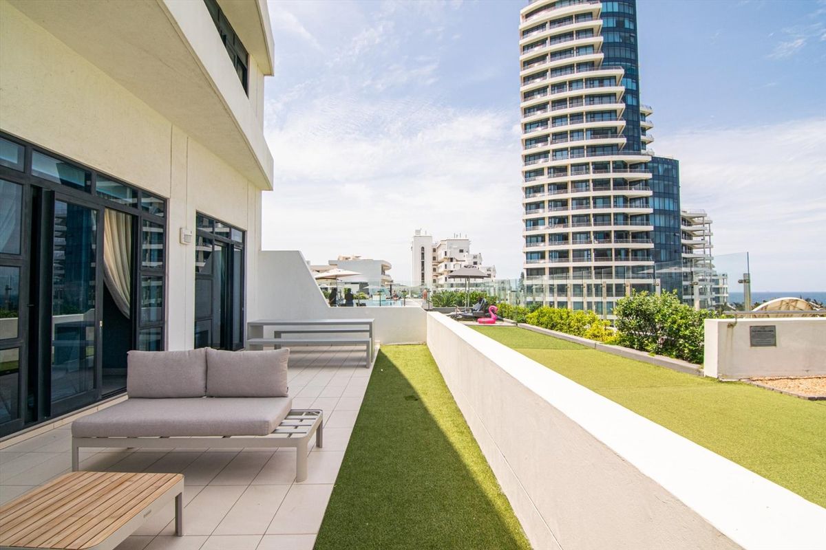 Spacious outdoor balcony featuring tiled flooring, artificial turf, and a modern glass balustrade with views of the surrounding high-rise buildings.