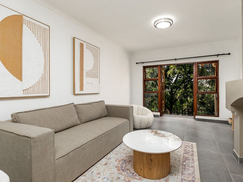 A bright living room featuring tiled flooring, a built-in fireplace, and wooden framed glass doors that open to a leafy outdoor view.