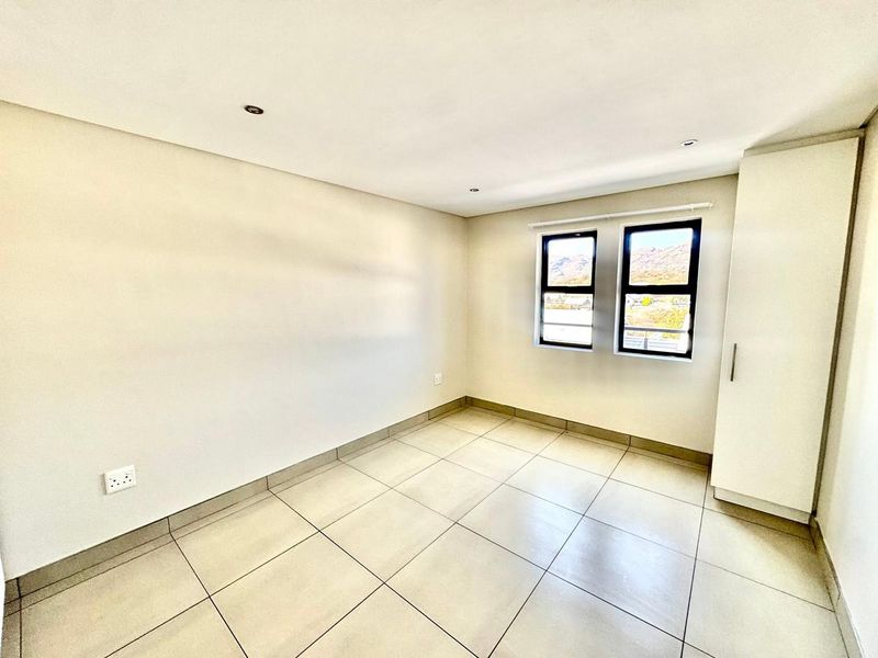A bright, empty bedroom featuring neutral tiled flooring, recessed downlighting, and a built-in wardrobe with views through the aluminium-framed windows.