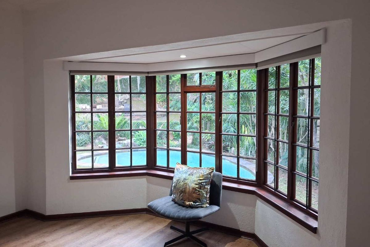 A bright living area featuring a large bay window with timber frames, offering a pleasant view of the garden and swimming pool.