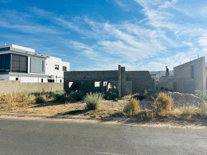 A vacant residential plot featuring an incomplete brick structure, perimeter walling with electric fencing, and unlandscaped ground in a developing neighborhood.