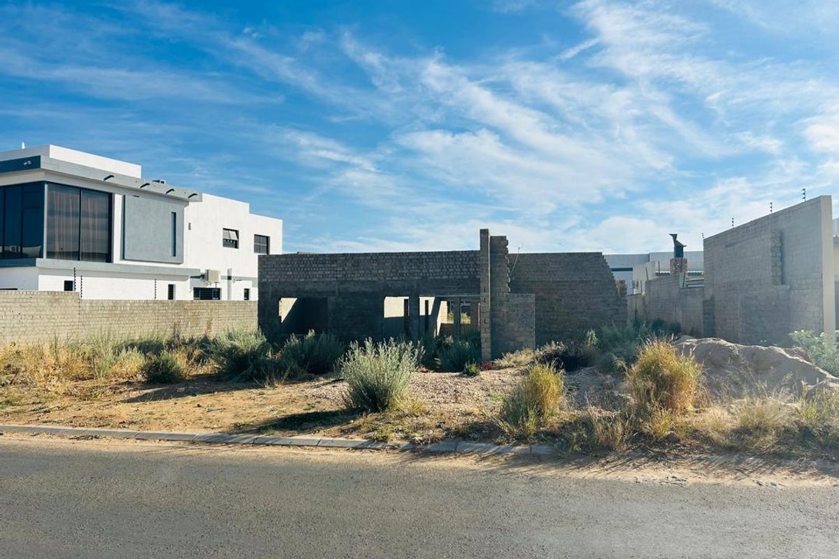 A vacant residential plot featuring an incomplete brick structure, perimeter walling with electric fencing, and unlandscaped ground in a developing neighborhood.