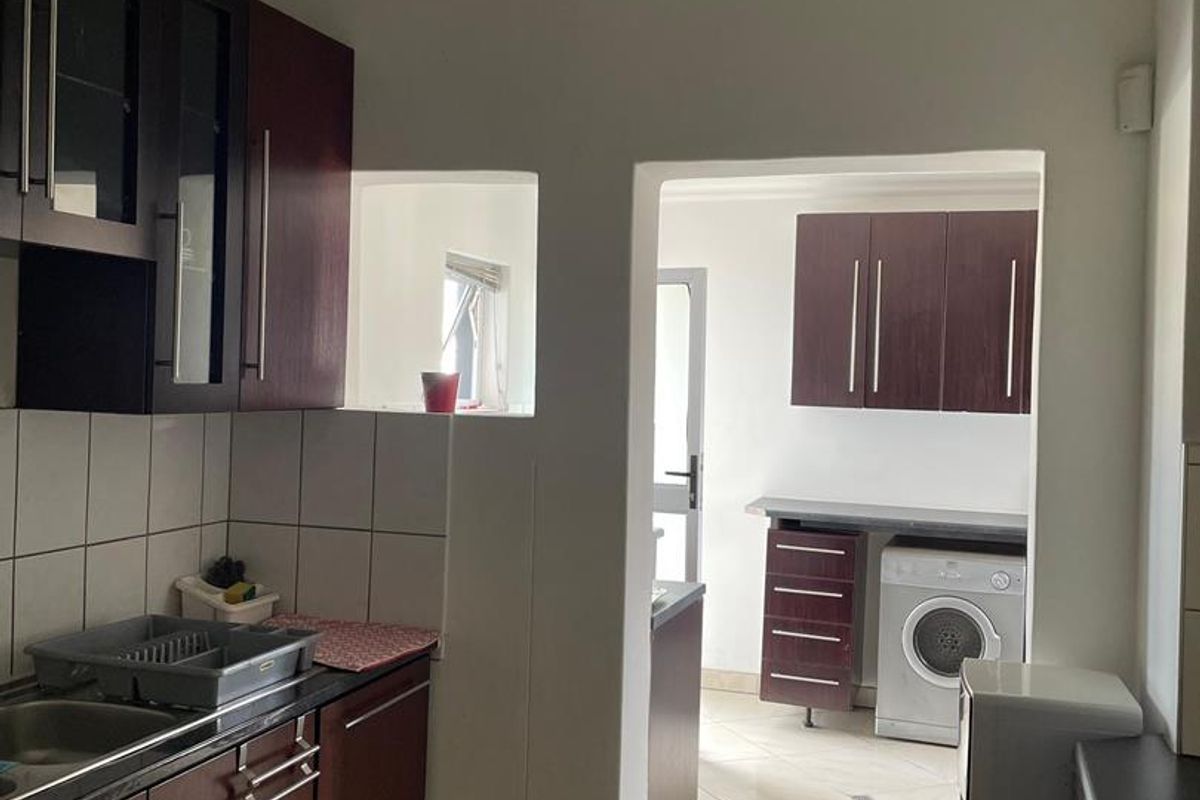 A functional kitchen featuring dark wood cabinetry, tiled backsplash, and laminate countertops, with an adjoining scullery area visible through the doorway.