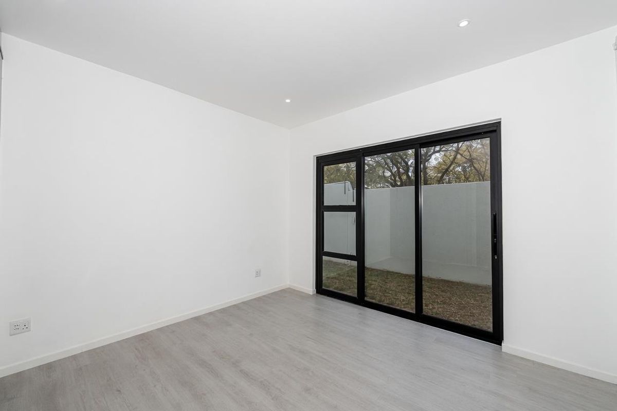 A bright, empty bedroom featuring modern light-toned laminate flooring, recessed ceiling lighting, and large black-framed sliding glass doors leading to a private outdoor area.