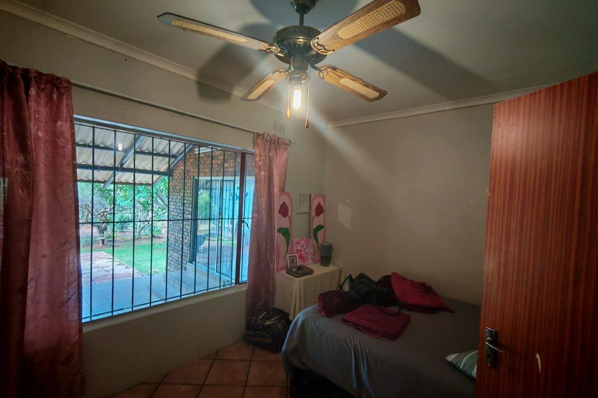A bedroom featuring terracotta floor tiles, a ceiling fan, and a large window with security bars overlooking the garden patio.