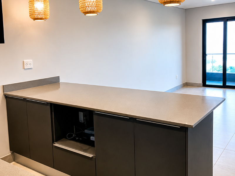 Modern kitchen island featuring dark cabinetry, stone countertops, and stylish pendant lighting, set within an open-plan living space with neutral tiled floors.