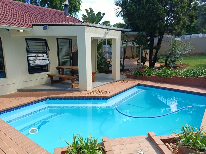 A well-maintained swimming pool area featuring a covered patio, brick paving, and established garden surroundings.