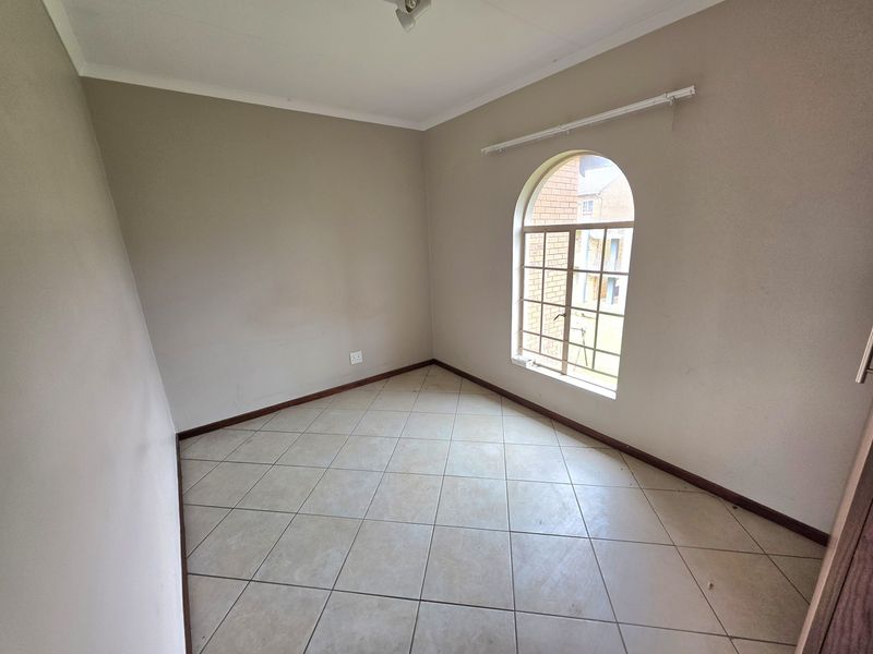 An empty bedroom featuring neutral tiled flooring, an arched window with security bars, and simple wall finishes.