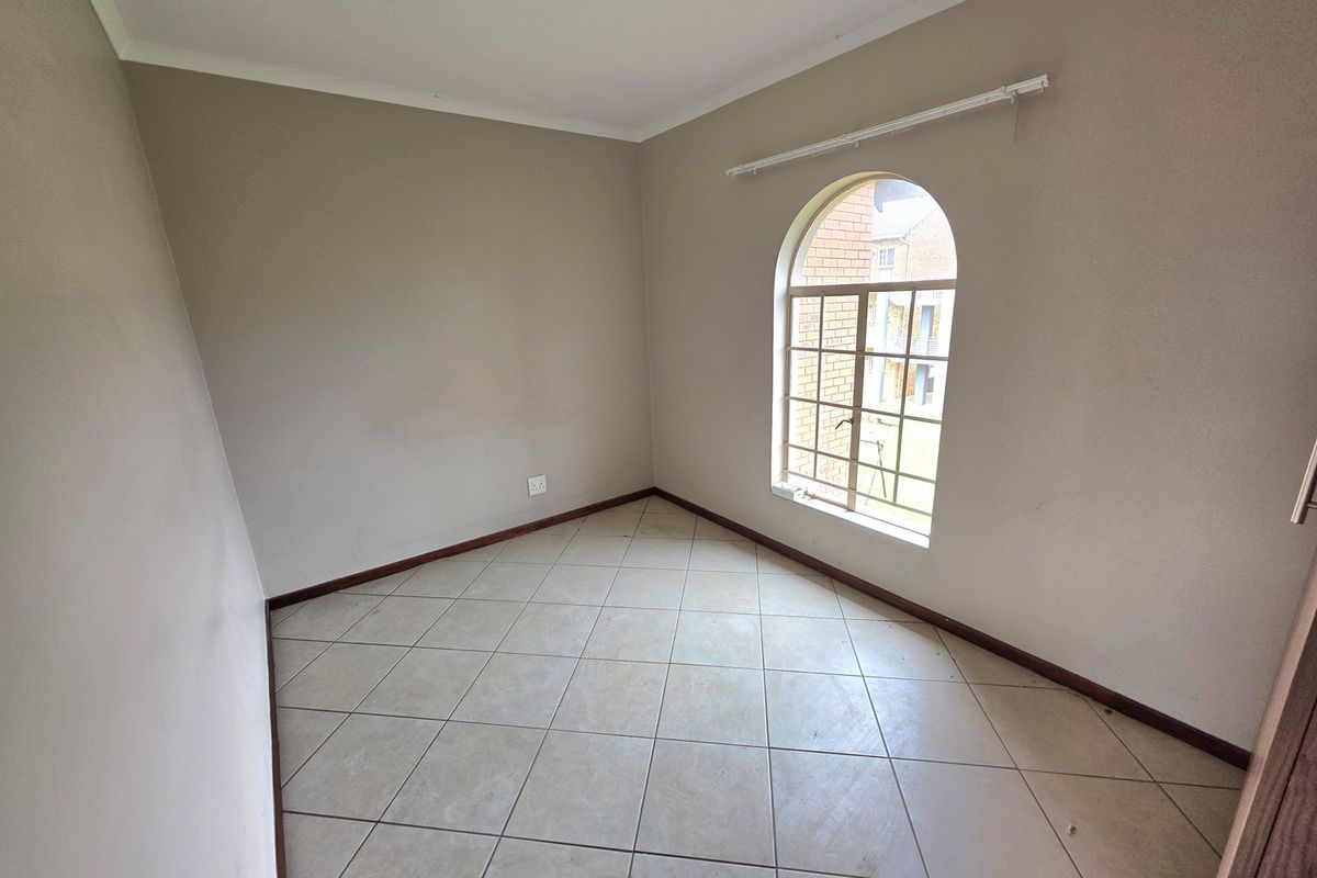 An empty bedroom featuring neutral tiled flooring, an arched window with security bars, and simple wall finishes.