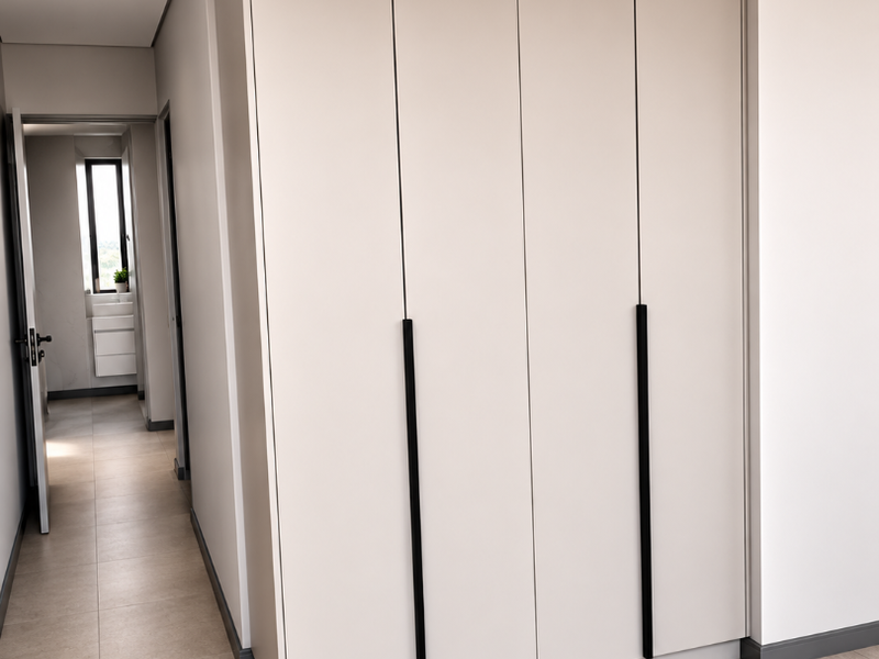 A modern bedroom featuring sleek, floor-to-ceiling built-in cupboards with matte black handles, neutral tiled flooring, and recessed downlighting.