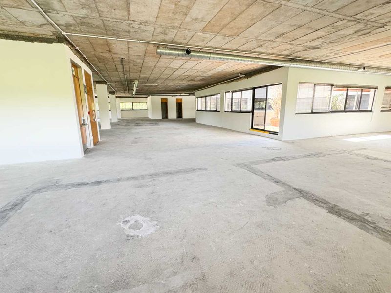 Spacious open-plan office area featuring an industrial aesthetic with exposed concrete ceilings, ducting, and large windows providing ample natural light.