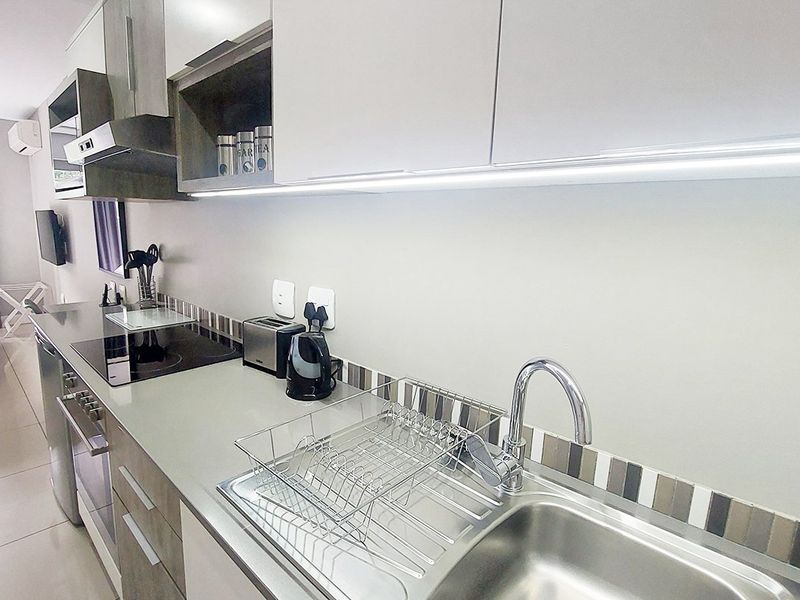 Modern galley-style kitchen featuring sleek cabinetry, integrated appliances, stainless steel sink, and contemporary tiled splashback, seamlessly flowing into the adjacent living area.