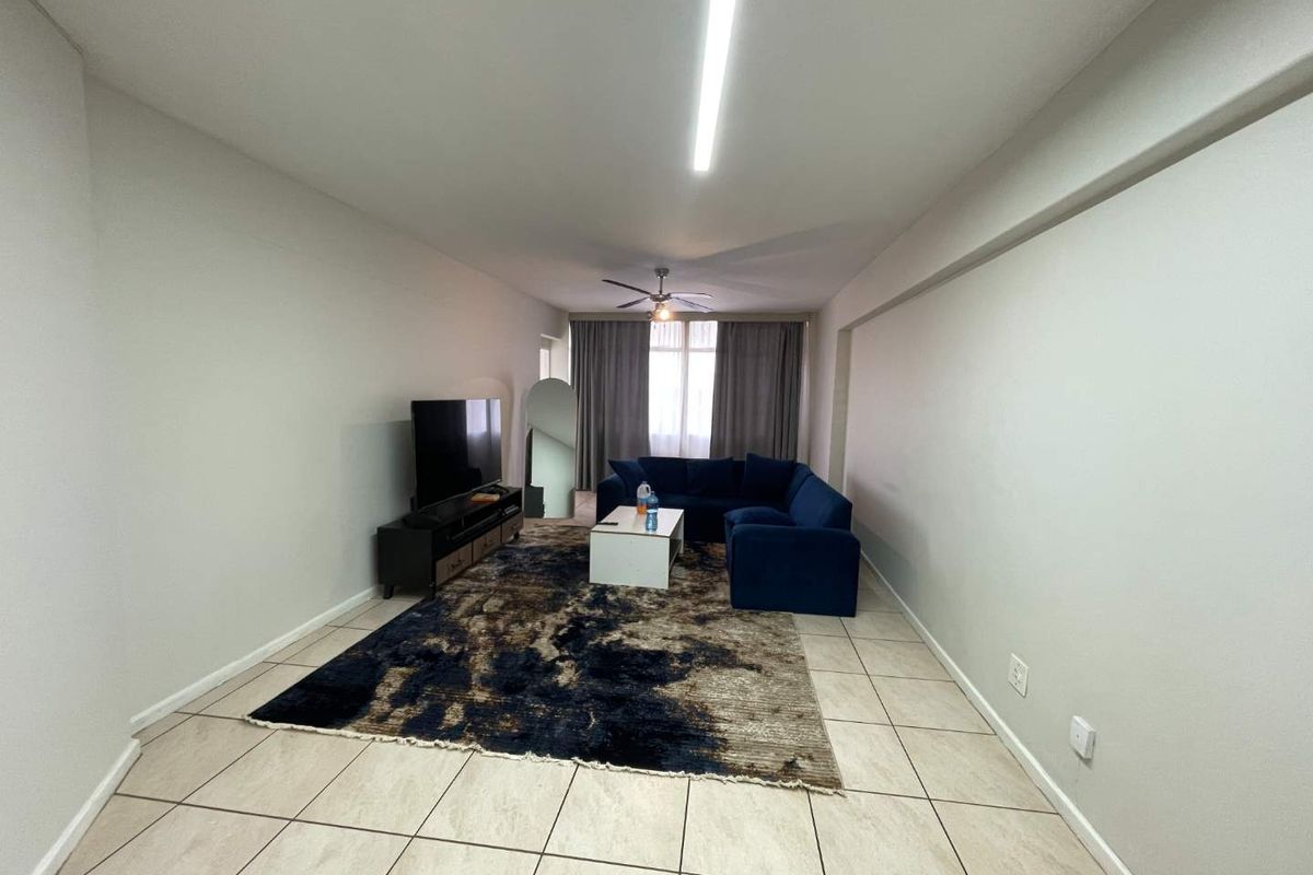 A spacious living area featuring neutral tiled flooring, a ceiling fan, and large windows with curtains, providing a bright and functional living space.